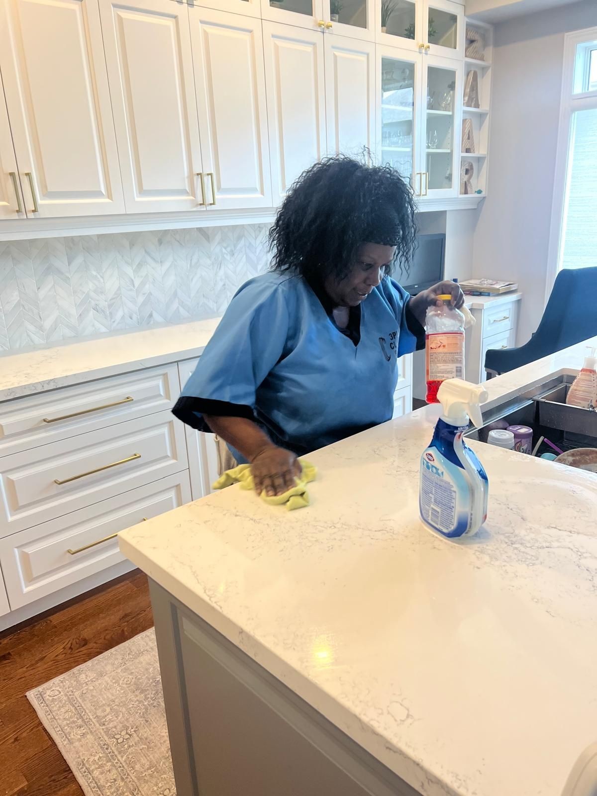 Person cleaning a kitchen counter with spray and cloth, surrounded by white cabinets and a bright window.